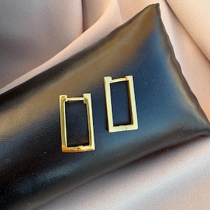 Peut inclure: Une paire de boucles d'oreilles créoles rectangulaires dorées. Les boucles d'oreilles sont présentées sur une surface en cuir noir, sur un fond doux aux tons neutres. Les boucles d'oreilles ont un design simple et moderne.