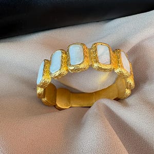 Può includere: Un bracciale rigido color oro con una finitura in metallo testurizzato e martellato, caratterizzato da cinque inserti rettangolari in pietra bianca iridescente disposti orizzontalmente lungo la parte anteriore. Il metallo ha un aspetto opaco e spazzolato.