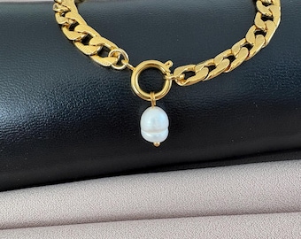 May include: A gold-colored chain bracelet with a large link design. The bracelet features a circular clasp and a single, white pearl pendant. The jewelry is displayed on a black surface with a neutral-toned fabric in the background.