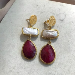 May include: A pair of gold-toned earrings with a layered design. Each earring features a hammered gold disc, a rectangular white pearl, and a teardrop-shaped, faceted, deep red gemstone. The gemstones are set in gold-toned bezels.