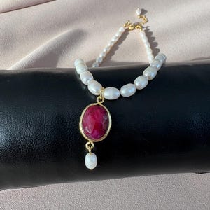 May include: A delicate bracelet featuring a strand of white, oval-shaped pearls. A gold-toned pendant with a large, oval-cut, red gemstone is the focal point. A smaller pearl dangles below the pendant, adding a touch of elegance.