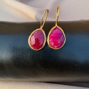 Könnte beinhalten: Ein Paar goldfarbene Ohrhänger. Jeder Ohrring hat einen tropfenförmigen, tiefroten Edelstein, der in eine strukturierte goldene Lünette gefasst ist. Die Ohrringe hängen an einem einfachen goldenen Haken.