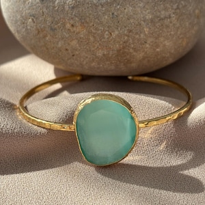 May include: A gold-toned cuff bracelet with a hammered texture and a large, oval, light-green gemstone. The bracelet is displayed on a neutral-colored fabric, with a large, gray stone in the background.