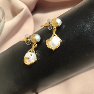 May include: A pair of gold-toned earrings with a pearl accent. The earrings have a textured design and a teardrop-shaped pearl pendant. The earrings are displayed on a black surface with a neutral-colored background.