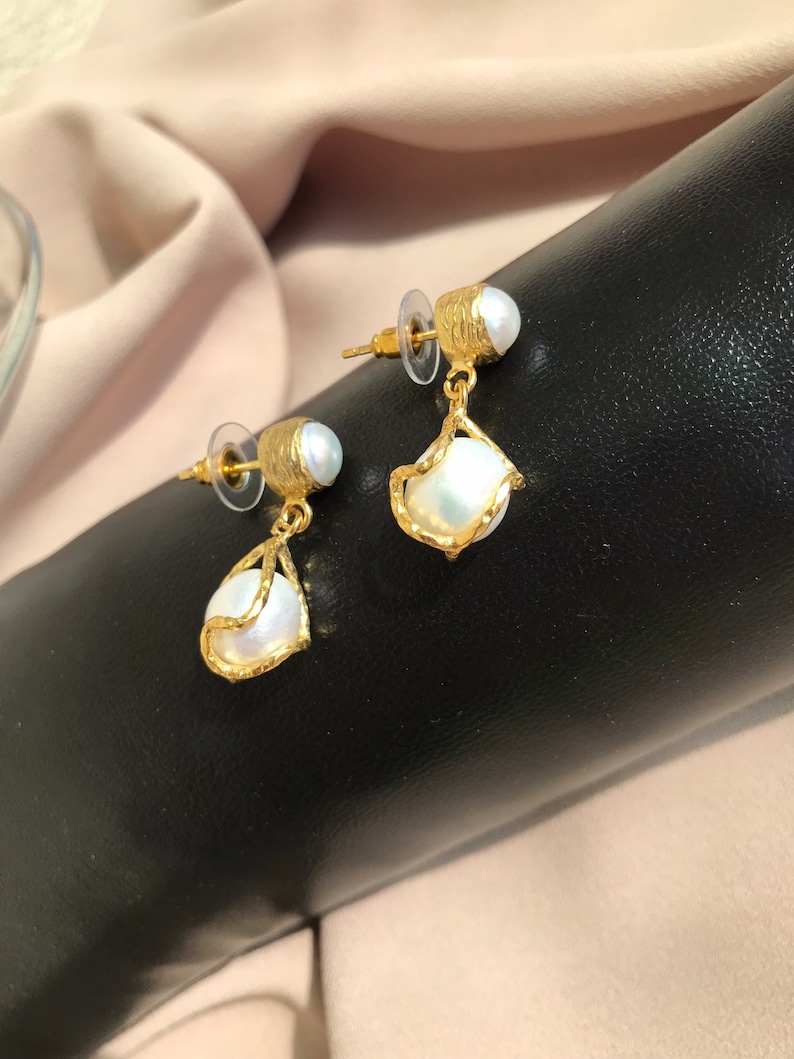 May include: A pair of gold-toned earrings with a pearl accent. The earrings have a textured design and a teardrop-shaped pearl pendant. The earrings are displayed on a black surface with a neutral-colored background.