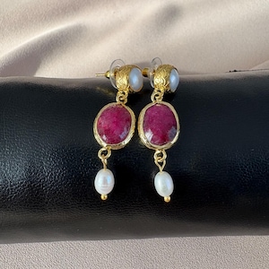 May include: A pair of gold-toned earrings featuring oval, deep red gemstones. Each earring has a small, white pearl at the top and a larger, teardrop-shaped pearl dangling below. The earrings are set against a black surface with a beige background.