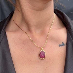Könnte beinhalten: Eine goldfarbene Halskette mit einem tropfenförmigen, rubinroten Anhänger. Der Anhänger ist goldgerahmt und hängt an einer zarten Goldkette. Die Halskette wird auf der Brust einer Person präsentiert.