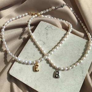 May include: Two pearl necklaces with initial pendants. One pendant is a gold letter "E" with a small diamond, and the other is a silver letter "B". The necklaces are on a marble surface.