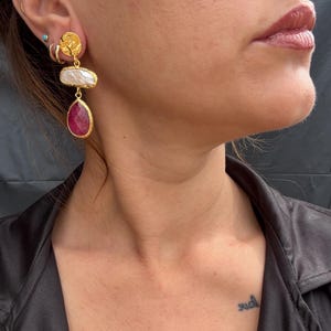 May include: Gold earrings featuring a textured disc, a white pearl, and a ruby-red gemstone. The earrings are suspended from the ear. The gemstones are set in gold. The earrings are worn on a person's ear.