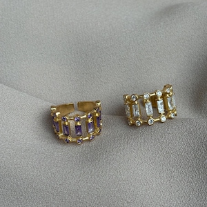 May include: Two gold-toned rings. One ring features rectangular purple stones and small round purple stones. The other ring has rectangular and round clear stones. Both rings are on a neutral background.
