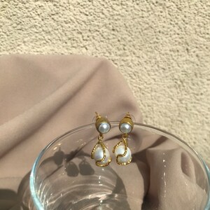 May include: A pair of gold-toned earrings with faux pearls. The earrings feature a small, round pearl at the top, with a larger, teardrop-shaped pearl suspended below, framed by a textured gold design. The background includes a neutral-toned fabric and a glass.