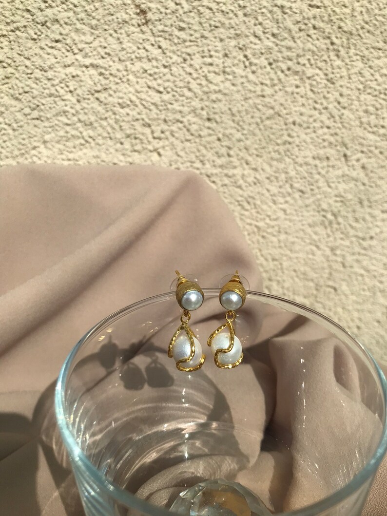 May include: A pair of gold-toned earrings with faux pearls. The earrings feature a small, round pearl at the top, with a larger, teardrop-shaped pearl suspended below, framed by a textured gold design. The background includes a neutral-toned fabric and a glass.