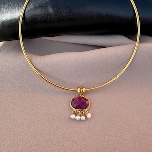 May include: A gold-toned choker necklace with a pendant featuring a large, oval-shaped, deep red gemstone. The pendant is accented with small, white pearls. The necklace is displayed on a neutral-toned surface.