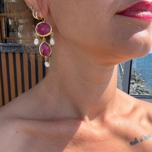 May include: A gold-toned dangle earring featuring two faceted, deep pink, oval-shaped stones set in gold bezels, accented with three small, irregular white freshwater pearls hanging from the bottom and sides of the stone settings.