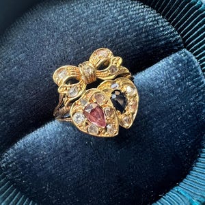 Op de afbeelding: Een gouden ring met een strik en twee hartvormige edelstenen. Eén hart is roze, de andere donkerblauw, beide omringd door heldere stenen. De ring ligt op een donkerblauwe fluwelen achtergrond.