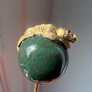 Op de afbeelding: Goudkleurige metalen speld met een gedetailleerde drakenfiguur bovenop een groene edelsteenbol. De draak heeft een robijnen oog. De edelsteen heeft rode vlekken. Een decoratief accessoire.