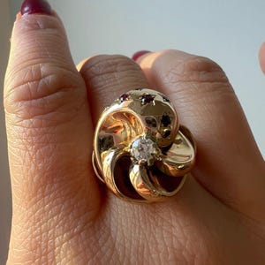 Könnte beinhalten: Ein goldfarbener Ring mit floralem Design. Der Ring hat einen zentralen, klaren Edelstein und ist mit kleineren roten Edelsteinen verziert. Das Design des Rings umfasst wirbelnde Blütenblätter und eine abgerundete Oberseite.