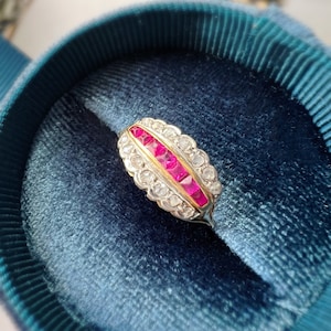 Peut inclure: Une bague en or avec une rangée de rubis rouges et une bordure de petits diamants. La bague est sertie sur une bande en métal blanc et est présentée dans un écrin à bague en velours bleu.