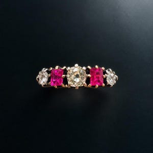 Könnte beinhalten: Ein Fünf-Stein-Ring mit einem zentralen Diamanten im Kissenschliff, flankiert von zwei rechteckigen, leuchtend pinken Rubinen und zwei äußeren Diamanten im Rundschliff, gefasst in eine Ringschiene aus Gelbgold mit silberfarbenen Krappen.