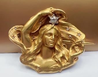 May include: A gold brooch featuring a woman's face with a diamond star on her head. The woman is holding the star with her right hand. The brooch is set with diamonds and has a flowing design.