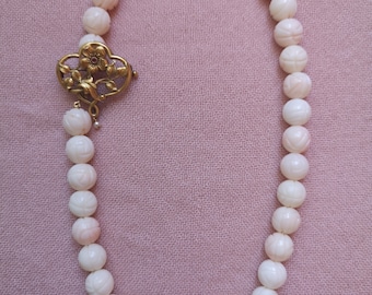 May include: A delicate necklace featuring carved, round, off-white beads with a floral design. The necklace is fastened with a gold-colored clasp with floral and leaf details. The background is a soft pink fabric.