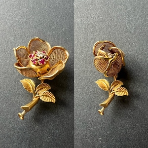 May include: A gold-toned rose brooch with a red and clear stone centre. The rose is made of delicate wire mesh and has a stem with leaves.