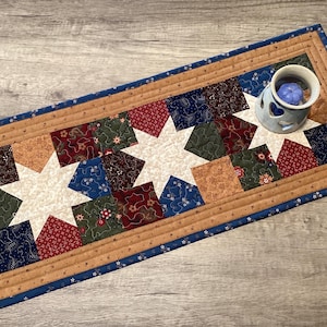 May include: A rectangular quilted table runner featuring three white star patterns surrounded by patchwork squares in shades of red, green, blue, and tan. The runner has a tan border with a thin blue trim and a small ceramic candle warmer.