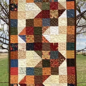 May include: A patchwork table runner with a brown border and a design of alternating triangles and squares in various shades of brown, red, orange, green, blue, and white.