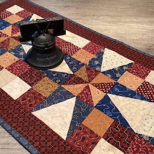May include: A patriotic table runner with a patchwork design in red, white, and blue. The runner features a variety of patterns and shapes, including squares, triangles, and floral prints. A black Liberty Bell statue sits on the runner.