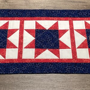 May include: A red, white and blue quilted table runner with a star pattern. The runner has a blue border with white stars and three red, white and blue star blocks in the centre. A small candle holder is sitting on the runner.