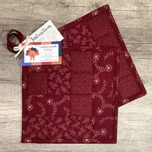 May include: Two square quilted potholders in a deep burgundy fabric featuring a white floral vine pattern and a small polka dot print. A white ribbon loop is attached to the corner, holding a tag labeled Instructions and Turkey Creek Boutique.