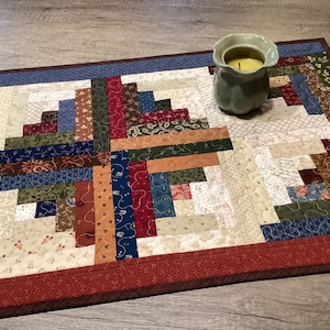May include: A rectangular fabric table runner featuring a log cabin quilt pattern with various printed cotton strips in shades of red, blue, green, and cream. The runner has a solid red border and a light green ceramic candle holder sits on top.