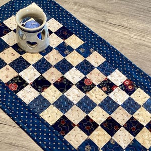 May include: A handcrafted quilted table runner with a blue and cream checkerboard pattern, accented with floral and polka dot details. A ceramic candle holder with heart cutouts sits on the runner, holding a blue candle.