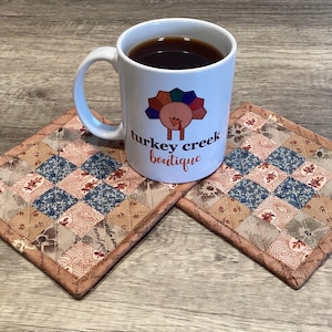 May include: A white ceramic mug filled with dark liquid, featuring a colorful turkey graphic and the text turkey creek boutique, resting on two square fabric coasters with a quilted patchwork pattern in shades of blue, tan, and brown.