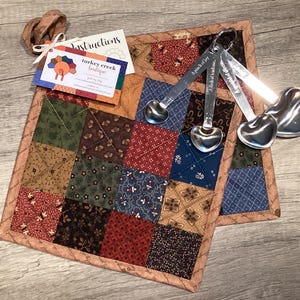 May include: A square quilted potholder featuring a patchwork pattern of various floral and geometric fabrics in shades of red, blue, brown, and green. A set of four heart-shaped stainless steel measuring spoons is attached, along with a branded tag.