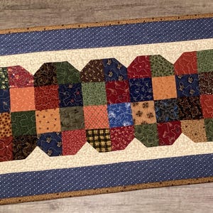 May include: A rectangular quilted table runner featuring a central patchwork design of multi-colored fabric squares in shades of red, green, blue, and brown, surrounded by a cream border and a blue polka-dot frame with a brown outer edge.