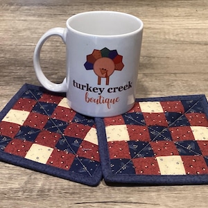 May include: A white ceramic mug with a turkey graphic and the text "turkey creek boutique" sits on two square quilted coasters. The coasters feature a patchwork design in red, white, and blue fabrics, with a dark blue border.
