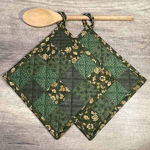 May include: Two square quilted potholders featuring a patchwork design of dark green, olive, and cream floral and geometric patterns, each with a fabric hanging loop at the top corner, positioned behind a light-colored wooden kitchen spoon.