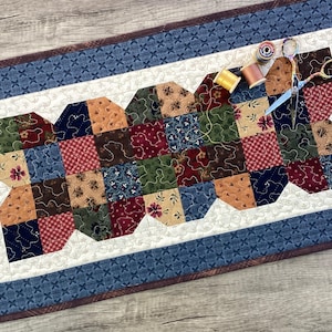 May include: A handmade patchwork table runner with a blue and white patterned border and a central design of various fabric squares in shades of red, green, and brown. Sewing supplies, including scissors and spools of thread, are placed on top.