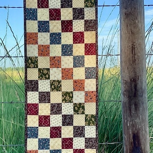 May include: A handmade patchwork table runner with a checkerboard pattern of squares in various colors, including burgundy, green, cream, and blue. The runner is hanging on a barbed wire fence with a wooden post in the background. The sky is blue.