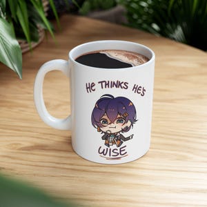 May include: White ceramic mug with a handle, filled with dark liquid. The mug features a cartoon illustration of a character with purple hair and the text "He thinks he's wise". The mug is on a wooden surface.