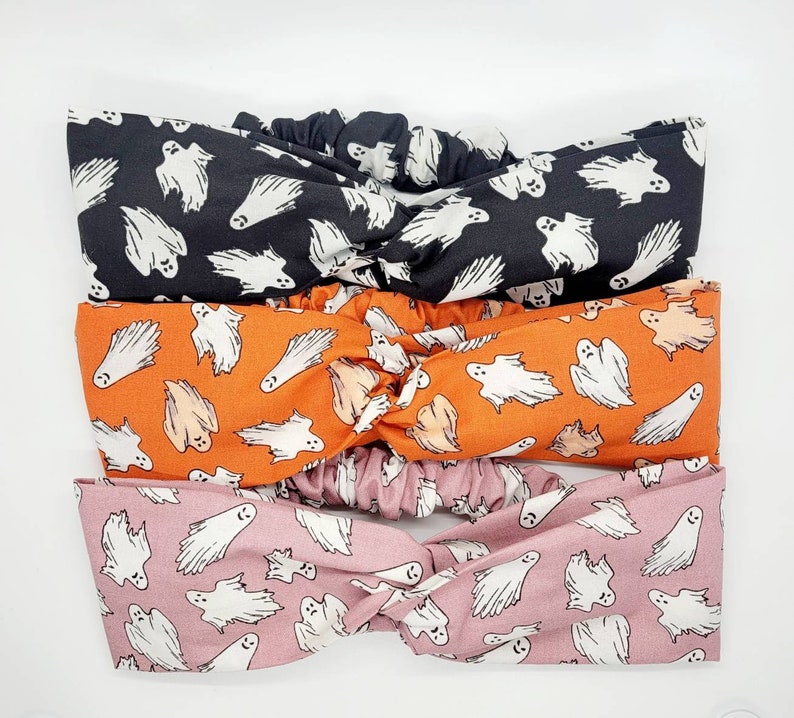 May include: Three fabric headbands with a knotted design. The headbands are printed with a pattern of white ghosts on a black, orange, and pink background.