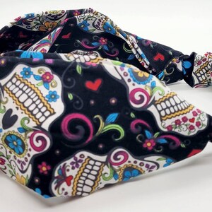 May include: Black headband with a colorful sugar skull pattern. The headband is made of a soft, stretchy fabric and has a knotted design.