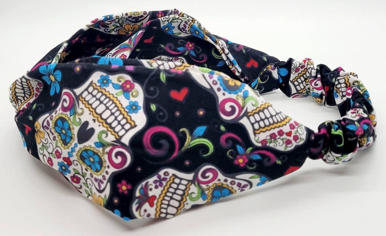 May include: Black headband with a colorful sugar skull pattern. The headband is made of a soft, stretchy fabric and has a knotted design.