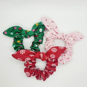 May include: Three Christmas-themed scrunchies with different patterns. One is green with lightbulbs, one is pink with white reindeer, and one is red with candy canes.