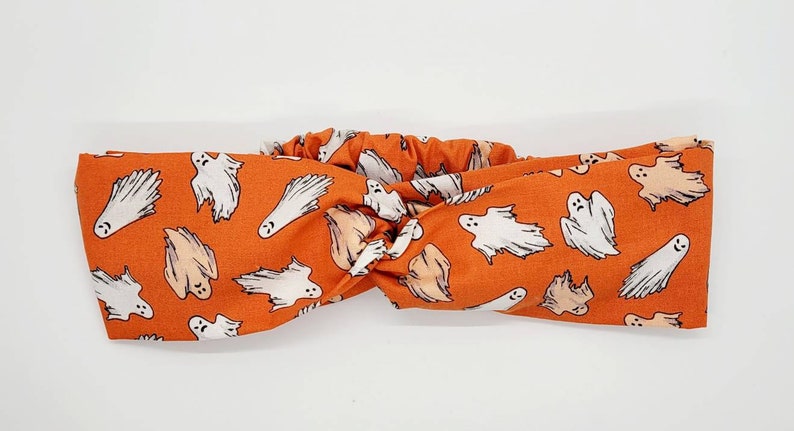 May include: Orange headband with a white ghost pattern. The headband is made of a stretchy fabric and is designed to be worn around the head.