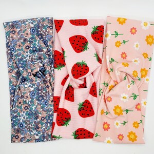 May include: Three fabric headbands with different patterns. One headband has a pink background with red strawberries, another has a pink background with yellow smiley faces, and the last one has a blue and white floral pattern.