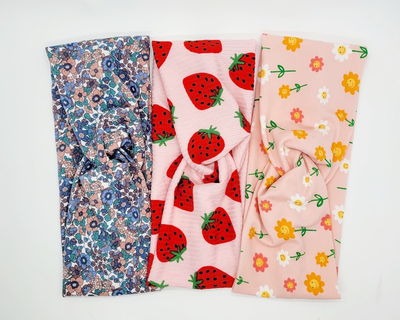 May include: Three fabric headbands with different patterns. One headband has a pink background with red strawberries, another has a pink background with yellow smiley faces, and the last one has a blue and white floral pattern.