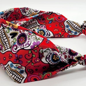 May include: Red headband with a white and colorful sugar skull pattern. The headband is made of a soft, stretchy fabric and has a knotted design.