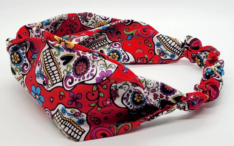 May include: Red headband with a white and colorful sugar skull pattern. The headband is made of a soft, stretchy fabric and has a knotted design.